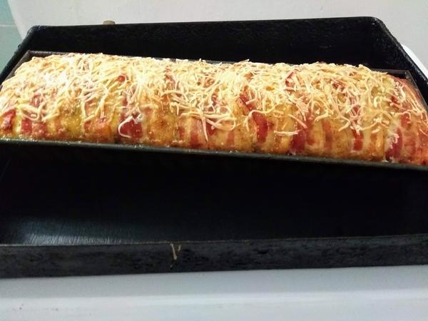 A húsos masszát belekanalazzuk a formába és ráhajtjuk a lelógó bacont. Hosszában a tetejére is tegyünk baconcsíkokat, ha nem fedné be teljesen a bacon a masszát.
Kenjük meg a félre tett tejföllel, majd szórjuk meg a maradék reszelt sajttal. Ha esetleg nem fér a massza a formába, süssük meg serpenyőbe, vagy tegyük papíros tepsibe a forma mellé sülni.